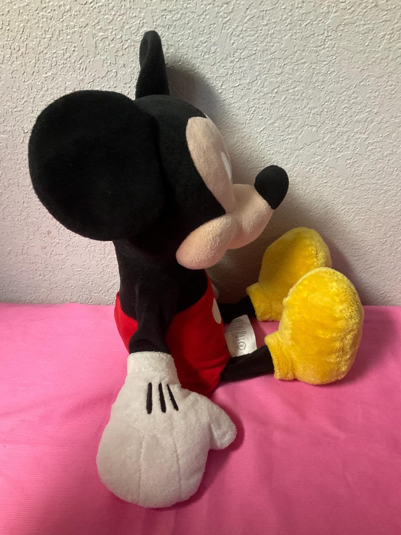May include: A plush Mickey Mouse toy, featuring a black head and ears, a white face, and a black nose. The toy wears red shorts, white gloves, and yellow shoes. The stuffed animal is sitting on a pink surface.
