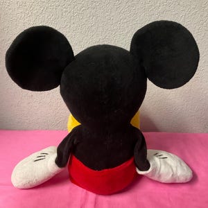 May include: A plush Mickey Mouse doll, viewed from the back. The doll features large black ears, a black head and torso, white gloves, and red shorts. The doll is sitting on a pink surface.