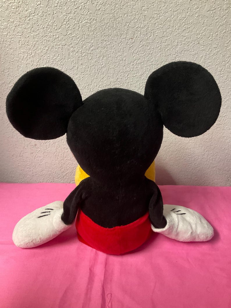 May include: A plush Mickey Mouse doll, viewed from the back. The doll features large black ears, a black head and torso, white gloves, and red shorts. The doll is sitting on a pink surface.