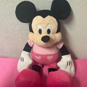 May include: A plush Mickey Mouse toy in a seated position. The toy features black ears, a tan face, and a pink shirt and shorts with heart accents. The hands are white with black lines, and the feet are large and pink.