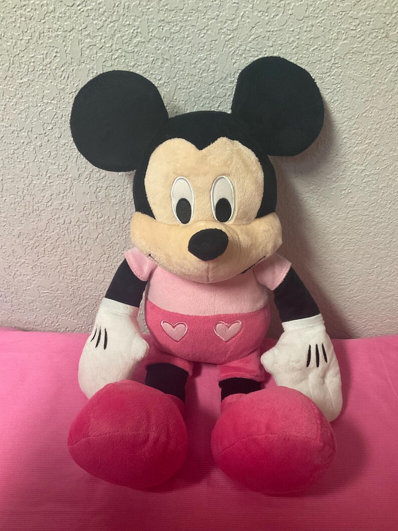 May include: A plush Mickey Mouse toy in a seated position. The toy features black ears, a tan face, and a pink shirt and shorts with heart accents. The hands are white with black lines, and the feet are large and pink.