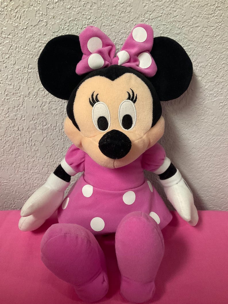 May include: A Minnie Mouse soft toy, with a pink dress and white polka dots, and a matching bow. The toy has black ears, a black nose, and white gloves. The toy is sitting on a pink surface.