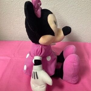 May include: A plush Minnie Mouse doll in a seated position. The doll has a black head and ears, a pink bow, and a pink dress with white polka dots. Her white gloves have black lines, and her pink shoes are visible.