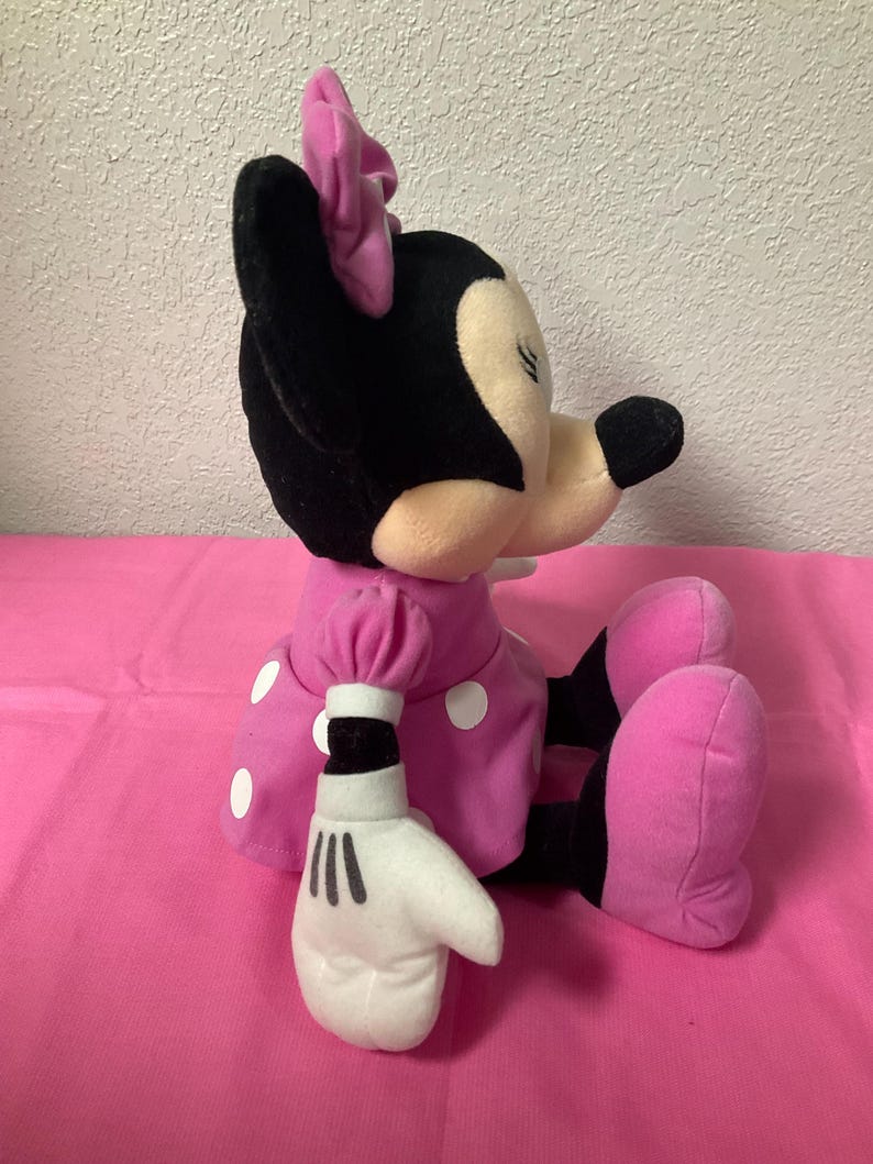 May include: A plush Minnie Mouse doll in a seated position. The doll has a black head and ears, a pink bow, and a pink dress with white polka dots. Her white gloves have black lines, and her pink shoes are visible.