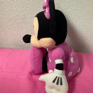 May include: A plush Minnie Mouse doll, with a black head and ears, a pink bow, and a pink dress with white polka dots. The doll's white gloved hand is extended, with black lines for fingers.