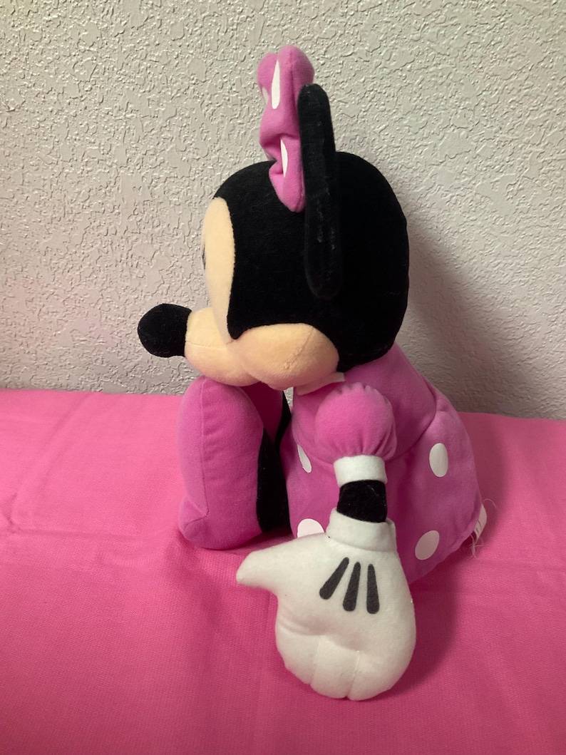 May include: A plush Minnie Mouse doll, with a black head and ears, a pink bow, and a pink dress with white polka dots. The doll's white gloved hand is extended, with black lines for fingers.