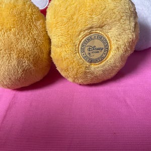 May include: Close-up of a yellow plush toy foot with a "Genuine Original Disney Store Authentic" patch. The foot is part of a larger toy, with red and white elements visible in the background. The toy rests on a pink surface.