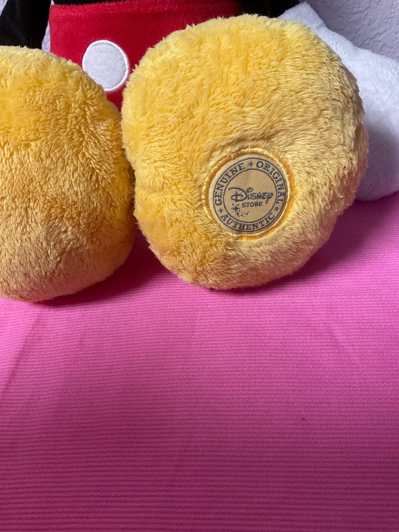 May include: Close-up of a yellow plush toy foot with a "Genuine Original Disney Store Authentic" patch. The foot is part of a larger toy, with red and white elements visible in the background. The toy rests on a pink surface.