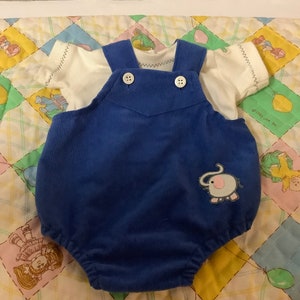 Op de afbeelding: Een blauwe corduroy baby romper met een olifant applique. De romper heeft witte bandjes en knopen. De romper ligt op een wit en geel dekbed.
