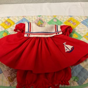 May include: A red sailor dress with a white collar and blue trim. The dress has a sailboat embroidered on the front and comes with matching red bloomers.