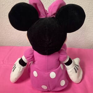 May include: A plush Minnie Mouse doll, seen from the back, with large black ears and a pink bow. The doll is dressed in a pink dress with white polka dots and white gloves. The background is a solid pink surface.