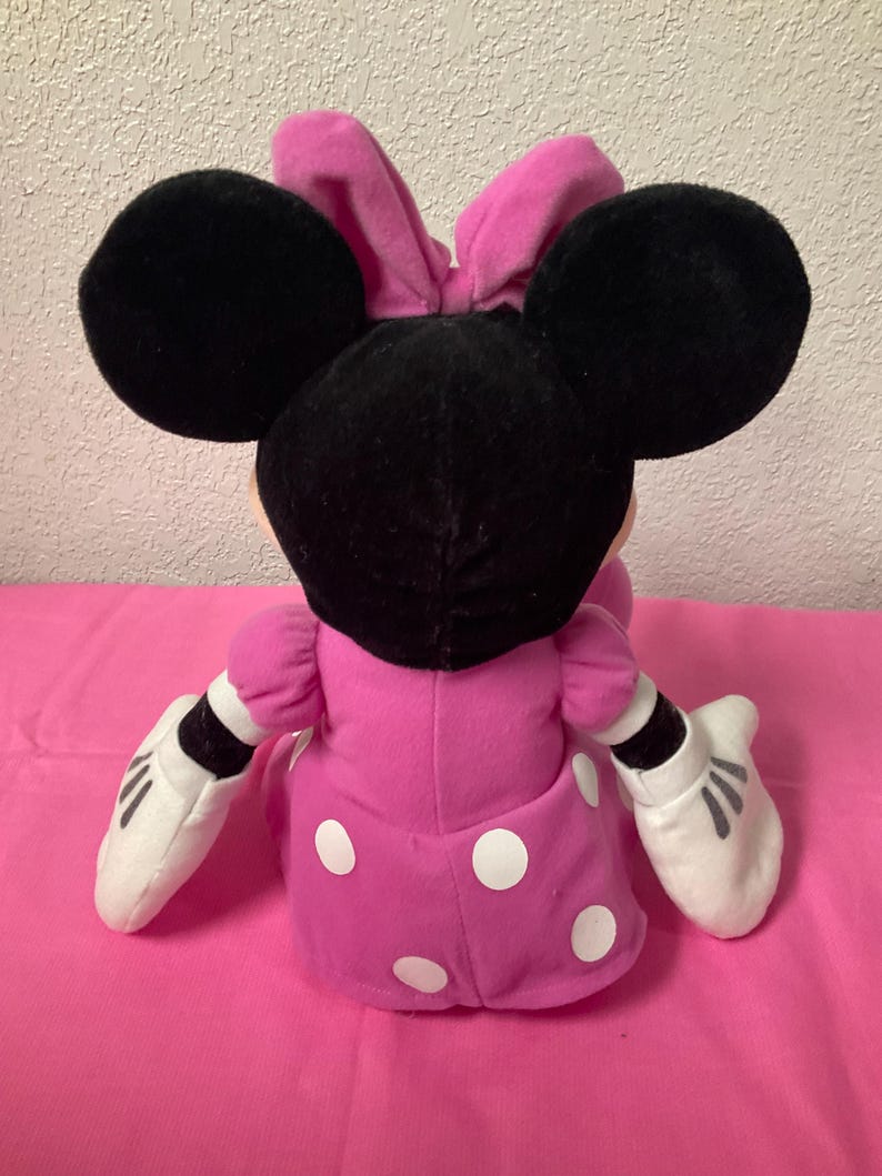 May include: A plush Minnie Mouse doll, seen from the back, with large black ears and a pink bow. The doll is dressed in a pink dress with white polka dots and white gloves. The background is a solid pink surface.