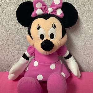 May include: A Minnie Mouse soft toy, with a pink dress and white polka dots, and a matching bow. The doll has black ears, a black nose, and white gloves. The toy is sitting on a pink surface.