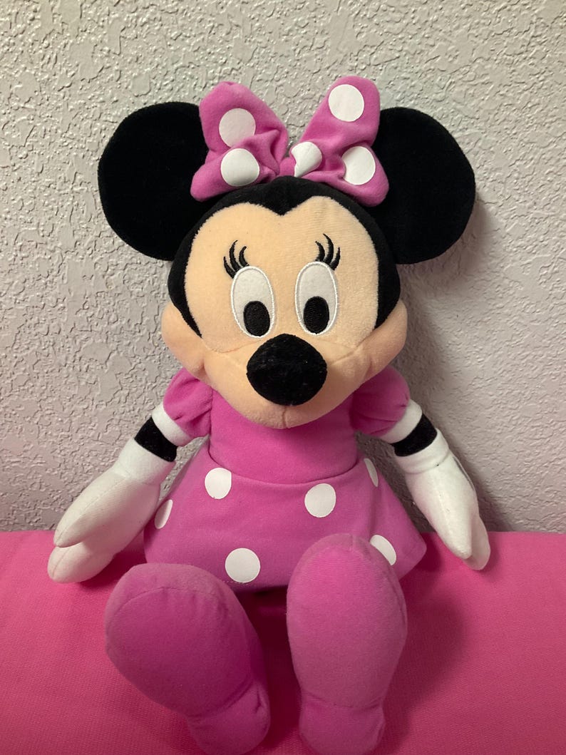 May include: A Minnie Mouse soft toy, with a pink dress and white polka dots, and a matching bow. The doll has black ears, a black nose, and white gloves. The toy is sitting on a pink surface.