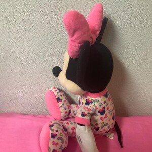 May include: A plush Minnie Mouse doll, featuring a black head, pink bow, and white gloves. The doll wears pajamas with a pattern of bows, stars, and rainbows. The doll is sitting on a pink surface.
