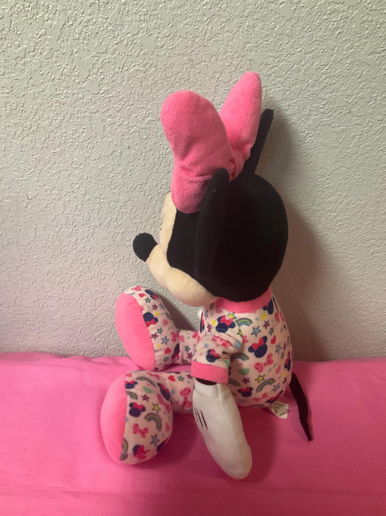 May include: A plush Minnie Mouse doll, featuring a black head, pink bow, and white gloves. The doll wears pajamas with a pattern of bows, stars, and rainbows. The doll is sitting on a pink surface.