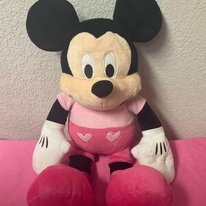 May include: A plush Mickey Mouse doll with black ears, a tan face, and a pink outfit. The doll has white gloves with black lines and large pink feet. Two pink hearts are on the front of the shorts.