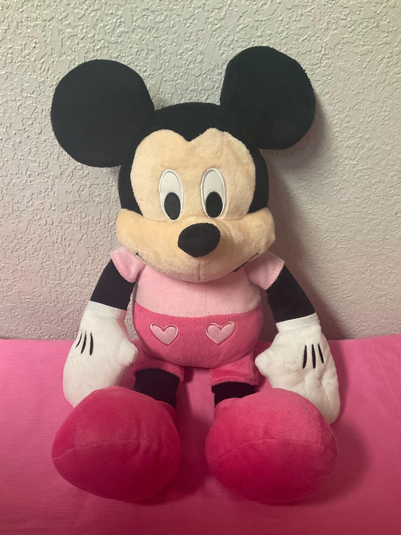 May include: A plush Mickey Mouse doll with black ears, a tan face, and a pink outfit. The doll has white gloves with black lines and large pink feet. Two pink hearts are on the front of the shorts.
