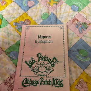 May include: A pink Cabbage Patch Kids adoption paper with green text and a cabbage graphic. The background is a quilted blanket with diamond patterns featuring Cabbage Patch Kids and balloons.