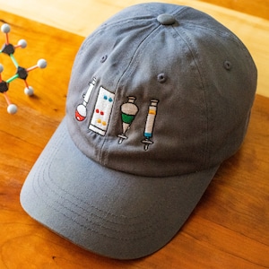 May include: Gray baseball cap with embroidered images of lab equipment, including a beaker, a flask, a graduated cylinder, and a syringe.