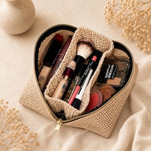 Könnte beinhalten: Eine beige gehäkelte Kosmetiktasche in Herzform mit goldenem Reißverschluss. Das Innere ist in zwei Fächer unterteilt und mit verschiedenen Kosmetika gefüllt, darunter eine Maybelline Master Brow Palette, Pinsel und Lippenstifte.