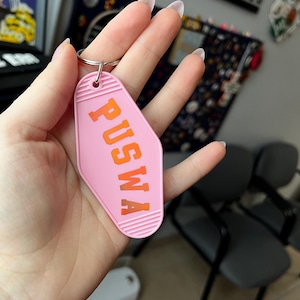 Può includere: Portachiavi rosa in stile motel con la scritta "PUSWA" in lettere arancioni. Il portachiavi è tenuto da un portachiavi argentato.