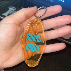 Puede incluir: Llavero naranja estilo motel con las palabras "BORING BARBIE" en turquesa. El llavero tiene un llavero plateado y está lleno de purpurina. El fondo está borroso.