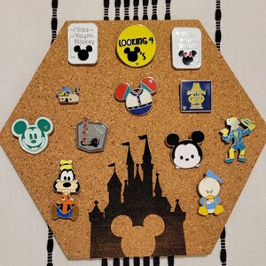 May include: A corkboard with a hexagon shape features a variety of Disney character pins. The pins are arranged in a pattern and include Mickey Mouse, Minnie Mouse, Goofy, Donald Duck, and other characters. The corkboard also features a silhouette of the Disney castle.