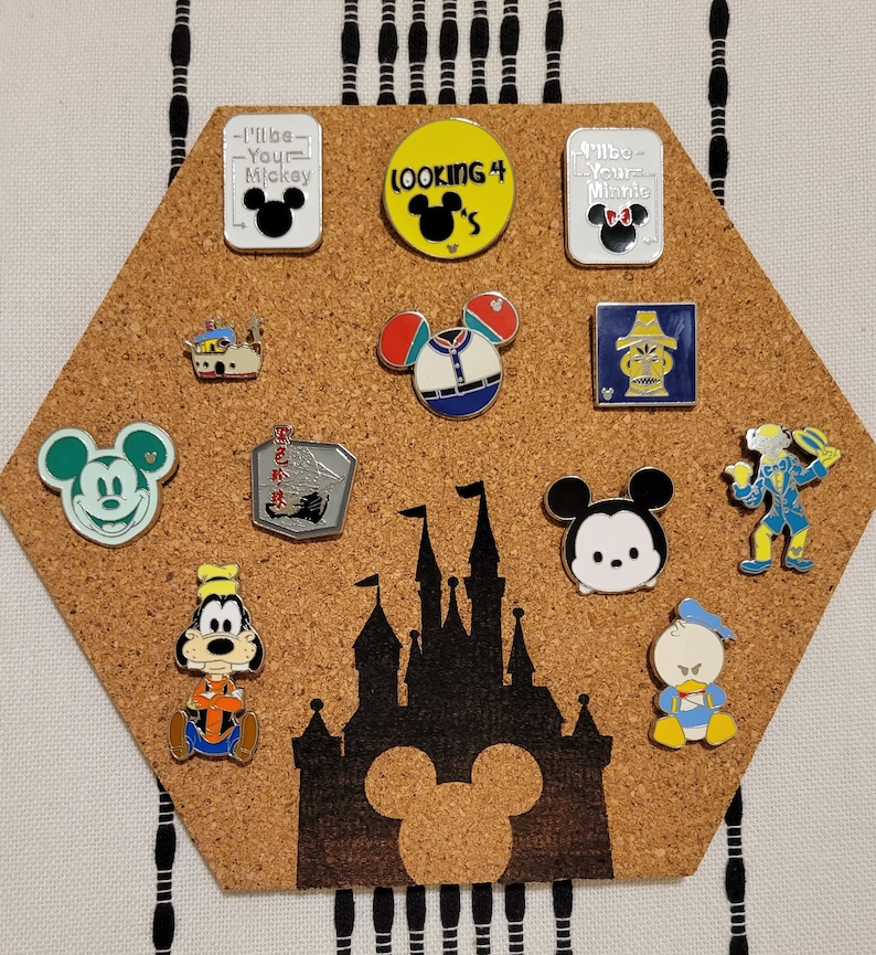 May include: A corkboard with a hexagon shape features a variety of Disney character pins. The pins are arranged in a pattern and include Mickey Mouse, Minnie Mouse, Goofy, Donald Duck, and other characters. The corkboard also features a silhouette of the Disney castle.