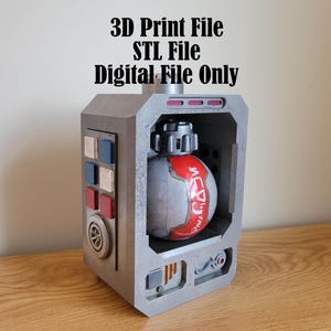 Puede incluir: Una carcasa rectangular gris impresa en 3D con un objeto esférico en su interior. La esfera es principalmente gris con detalles en rojo y texto japonés. La carcasa tiene botones y un diseño circular en el lateral. El texto superior dice: 3D Print File, STL File, Digital File Only.