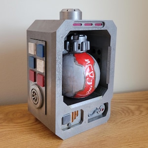 Op de afbeelding: Een grijze, rechthoekige doos met een bolvormig object erin. De bol is wit met rode accenten en Japanse tekst. De doos heeft knoppen in rood, wit en blauw en een cirkelvormig ontwerp. Het object staat op een houten oppervlak.