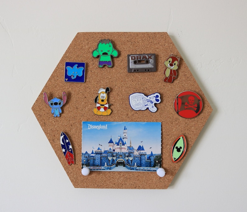 May include: A corkboard with a variety of pins, including a blue Stitch character, a green Hulk character, a red pirate skull and crossbones, a blue and white Mickey Mouse character, a yellow and red Donald Duck character, a blue and white Dumbo character, a red and white Chip character, a blue and white cassette tape, and a postcard with a picture of Sleeping Beauty's castle. The corkboard is hexagonal in shape.