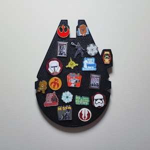 May include: A black Millennium Falcon-shaped pin board with various Star Wars themed pins attached. The pins feature characters, logos, and quotes from the Star Wars franchise.