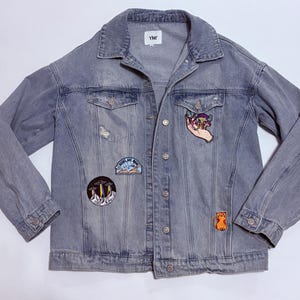 May include: A light blue denim jacket with multiple patches. The jacket has a classic collar, button closures, and two chest pockets. Patches include a hand holding mushrooms, a cat, and a UFO scene. The jacket has a distressed look with a small tear.