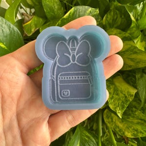 May include: A light blue silicone mold in the shape of a backpack with a bow and mouse ears. The mold has a detailed design of a backpack with a pocket and a small mouse head logo. The mold is held in a hand against a background of green leaves.
