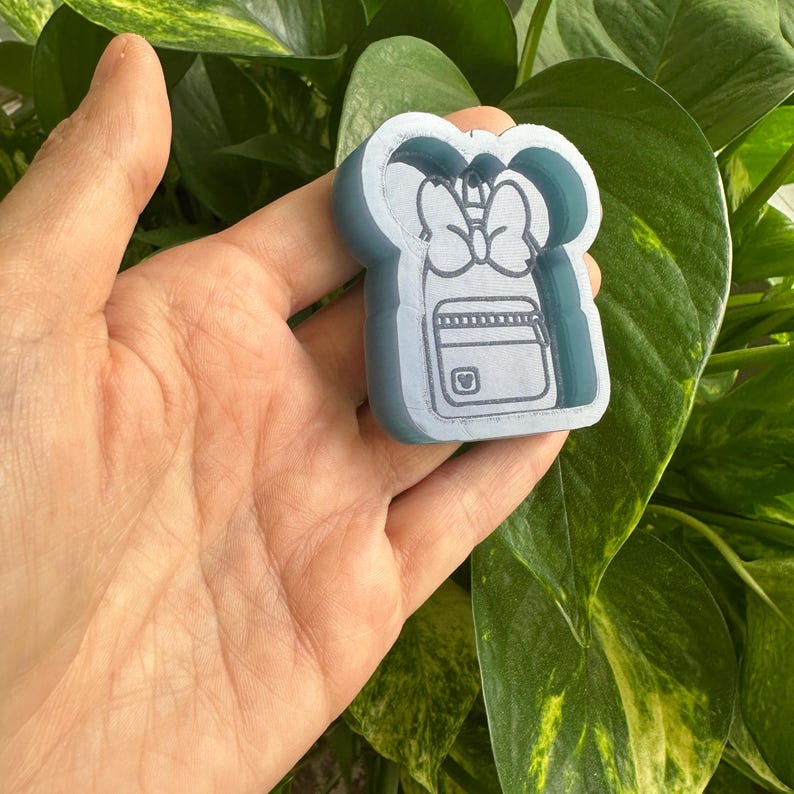 May include: A light blue, 3D-printed backpack-shaped object with a bow and a pocket. The object is held in a hand against a background of green leaves. The backpack design has a bow on top and a pocket with a small logo.