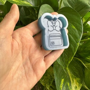 May include: A light blue, 3D-printed backpack-shaped object with a bow and a pocket. The object is held in a hand against a background of green leaves. The backpack design includes a bow, a pocket, and a small logo.