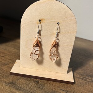 May include: A pair of earrings featuring clear quartz crystals wrapped in copper wire. The earrings are hanging from silver hooks on a wooden display stand.