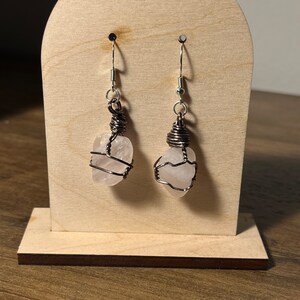 May include: A pair of earrings featuring clear quartz stones wrapped in copper wire. The earrings are suspended from silver hooks on a wooden display stand.