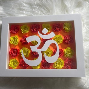 Puede incluir: Imagen enmarcada en blanco con un símbolo Om de papel blanco rodeado de flores de papel rojo y amarillo sobre un fondo naranja.