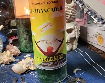 Puede incluir: Una vela amarilla y verde con el texto "VELADORA DE COCKTAIL DESTRANCADERA UNCHAINED" y una imagen de una persona liberándose de cadenas. La vela está sobre una superficie azul con una calavera y una cadena.