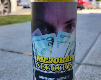 Puede incluir: Una vela de vidrio azul con una capa de cera amarilla y una etiqueta que dice "Mejorar Negocios" e "Improve Business". La etiqueta también presenta los ojos de una persona y una pila de billetes de veinte dólares estadounidenses.