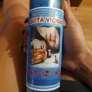 Puede incluir: Una vela de vidrio azul con la etiqueta "VELADORA DE COCKTAIL QUITA VICIOS" con una imagen de una persona y una botella. La vela tiene un interior de cera azul y se sostiene contra un fondo de piel y madera.