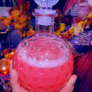 May include: A clear, round glass bottle with a textured surface, filled with a pink liquid and topped with a crystal stopper. The bottle is held in a hand, with a blurred background of flowers and other objects.