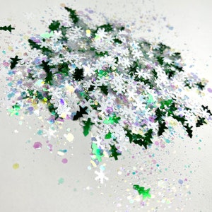 May include: A pile of iridescent green and white glitter in the shape of snowflakes and Christmas trees. The glitter is scattered on a white background.