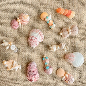 May include: Assortment of seashell hair clips with pearl and turquoise accents. The clips are arranged on a burlap background.