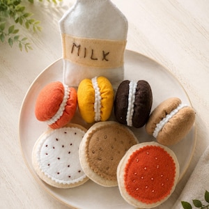 May include: A collection of felt play food items on a plate, including a white milk carton with the word MILK embroidered on it, four colorful sandwich cookies, and three flat round cookies in white, tan, and orange with stitched details.