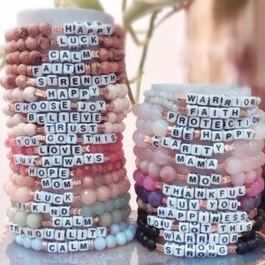 May include: A stack of colorful beaded bracelets with positive messages spelled out in black letters. The bracelets feature words like "Happy", "Faith", "Warrior", "Love", "Hope", "Calm", "Strength", "Trust", and "Mom".