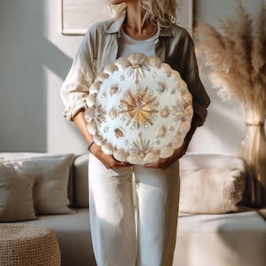May include: A round, white decorative piece adorned with various seashells, pearls, and a central starburst design. The edges are lined with scallop shells. The piece is held in front of a neutral background.
