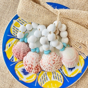 May include: A decorative plate with a blue and yellow pattern, topped with a beige burlap napkin and seashell napkin rings. The napkin rings feature white wooden beads, turquoise accents, and pink and white seashells.